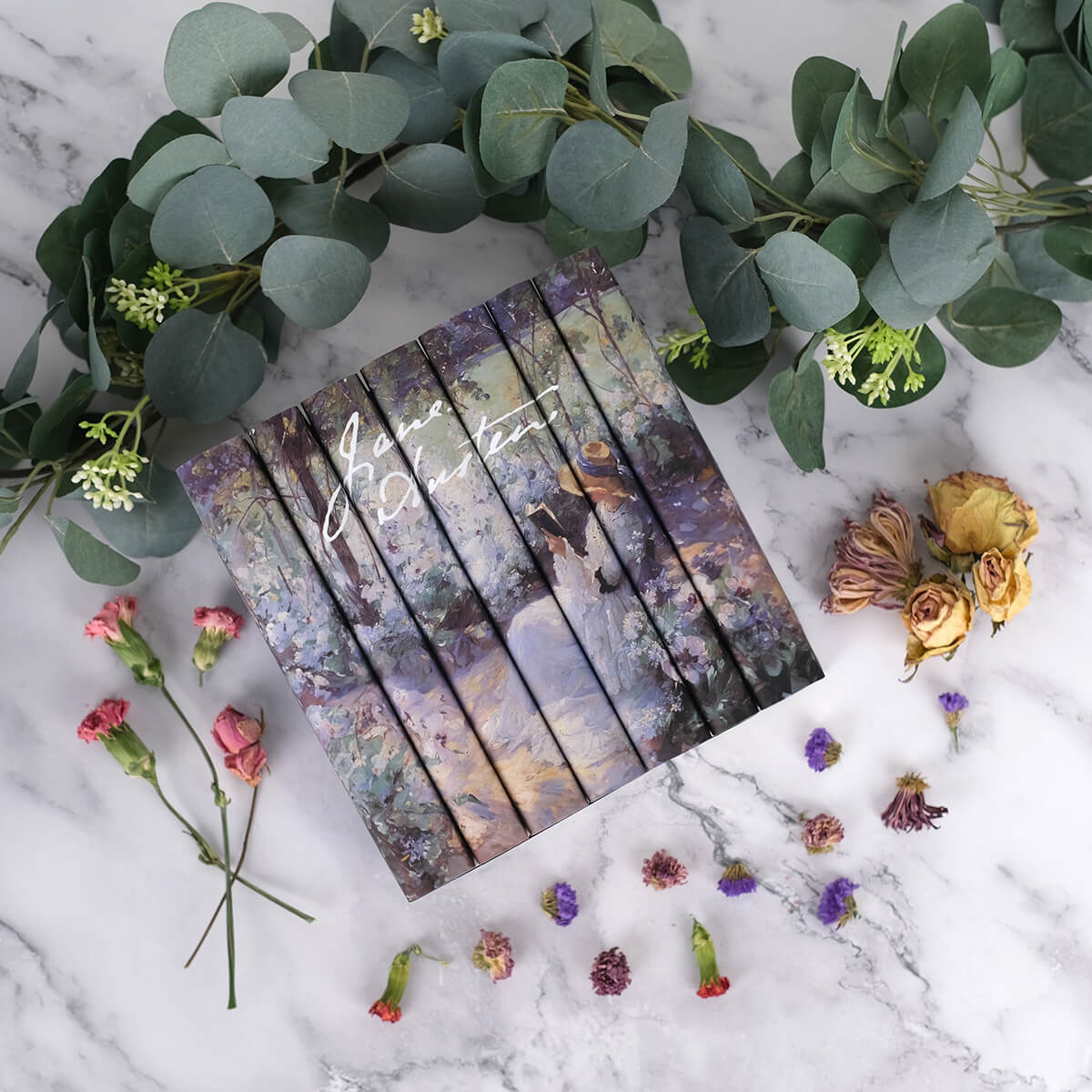 Jane Austen Delicious Solitude Book set from Juniper Custom surrounded by dried flowers and green leaves sitting against a backdrop of white marble.