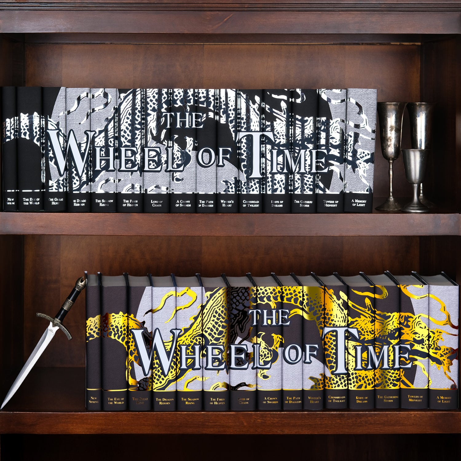 Two Wheel of Time Dragon Reborn Limited Edition Book Sets from Juniper Books sitting on a dark wooden bookshelf. The top shelf is designed in silver foil and the bottom in gold foil. The shelves are decorated with silver cups and a ornate dagger.