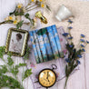 Flat-lay arrangement featuring a Brontë sisters book set with Monet-inspired dust jackets, showcasing 'Woman with a Parasol.' The setup includes vintage-inspired elements: a gold-framed portrait, a classic pocket watch, a teacup, handwritten script, floral accents, and greenery. The scene evokes a sense of timeless elegance and literary charm.