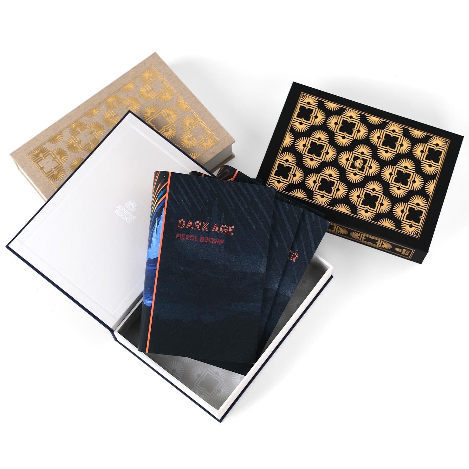 Three open dust jacket boxes designed by Juniper Books, shown from above. Each box displays an intricate, symmetrical pattern. The beige box has gold patterns, the black box has gold patterns, and the navy blue box has silver patterns. The interiors of the boxes feature a lighter version of the pattern and the Juniper Books logo on the inside cover of the navy blue box. A set of Pierce Brown Red Rising Extension Dust Jackets sit in the open box.