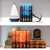 Percy Jackson and the Olympians book set on a white shelf sitting above the Heroes of Olympus book set surrounded by antique books, a sailboat, and a laurel crown.