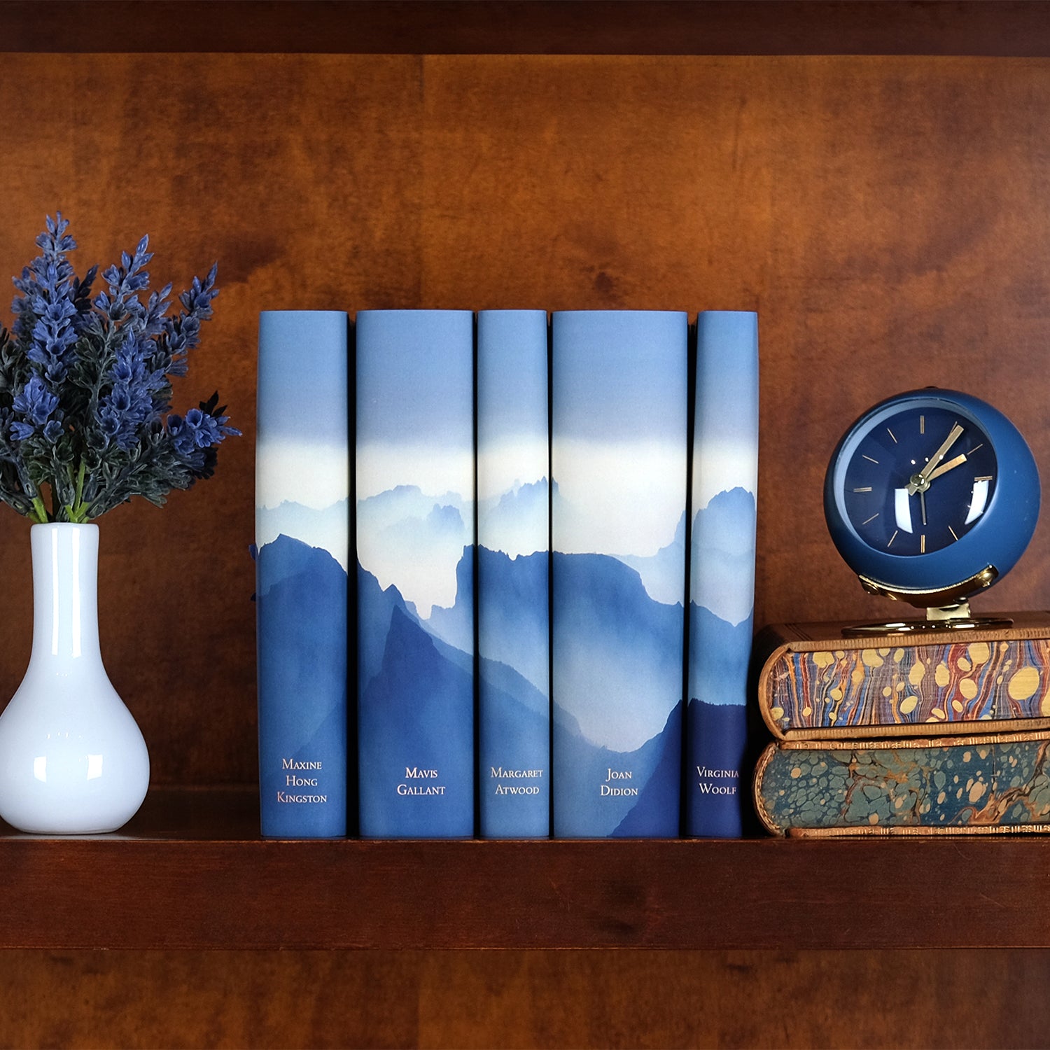 A wooden bookshelf is neatly arranged with decorative book sets and accessories. On the top shelf, a collection of five hardcover books with blue-toned mountain landscape dust jackets is displayed upright, alongside a small stack of vintage books with a blue clock on top and a wooden vase holding dried flowers. The bottom shelf showcases two sets of books with vibrant, artistic dust jackets One set features a bold, colorful portrait of Margaret Atwood and the other a colorful portrait of Maya Angelou.