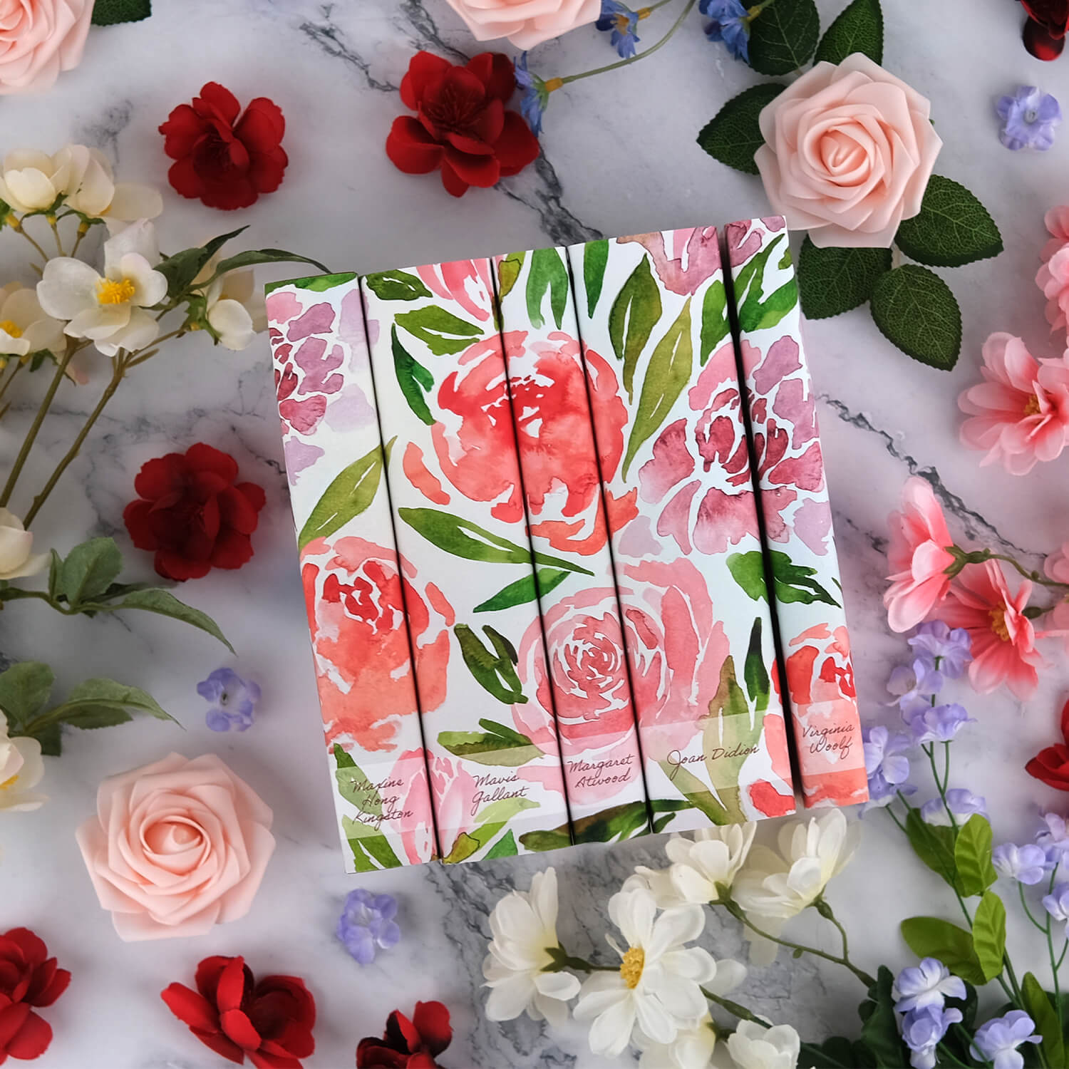 The Spring Time Influential Women Book Set from Juniper Custom sits against a white marble background surrounded by roses, blue cornflowers, white daisies, and white wild roses. 