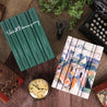 Green Hemingway Signature set and Essentials Ernest Hemingway Book Set sitting on a woven clothes against a wood background surrounded by antique leather books, a typewriter, plants, and a clock.