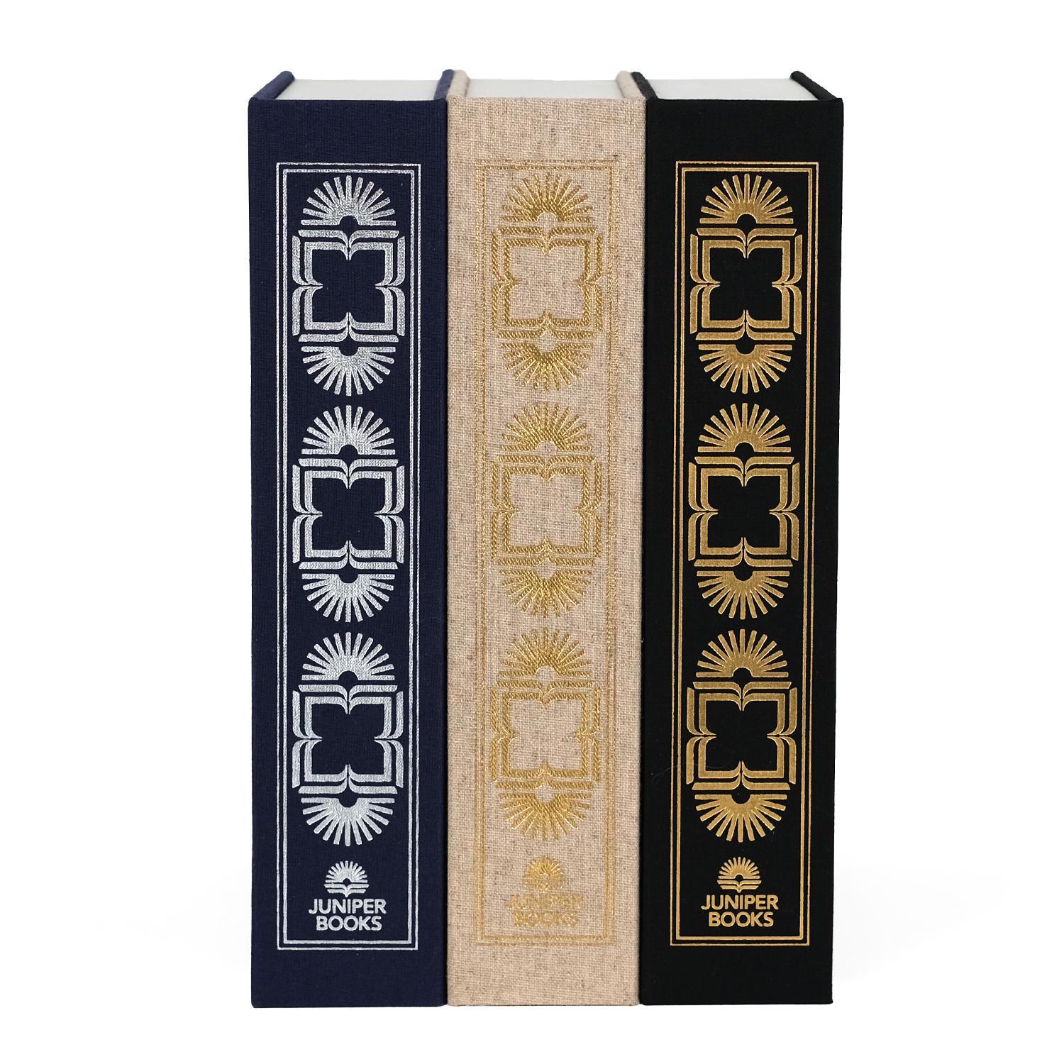 A set of three dust jacket boxes designed by Juniper Books, resembling hardcover book spines. Each box features intricate, symmetrical patterns in metallic colors—silver on a navy blue box, gold on a beige box, and gold on a black box. The Juniper Books logo is displayed at the bottom of each spine.